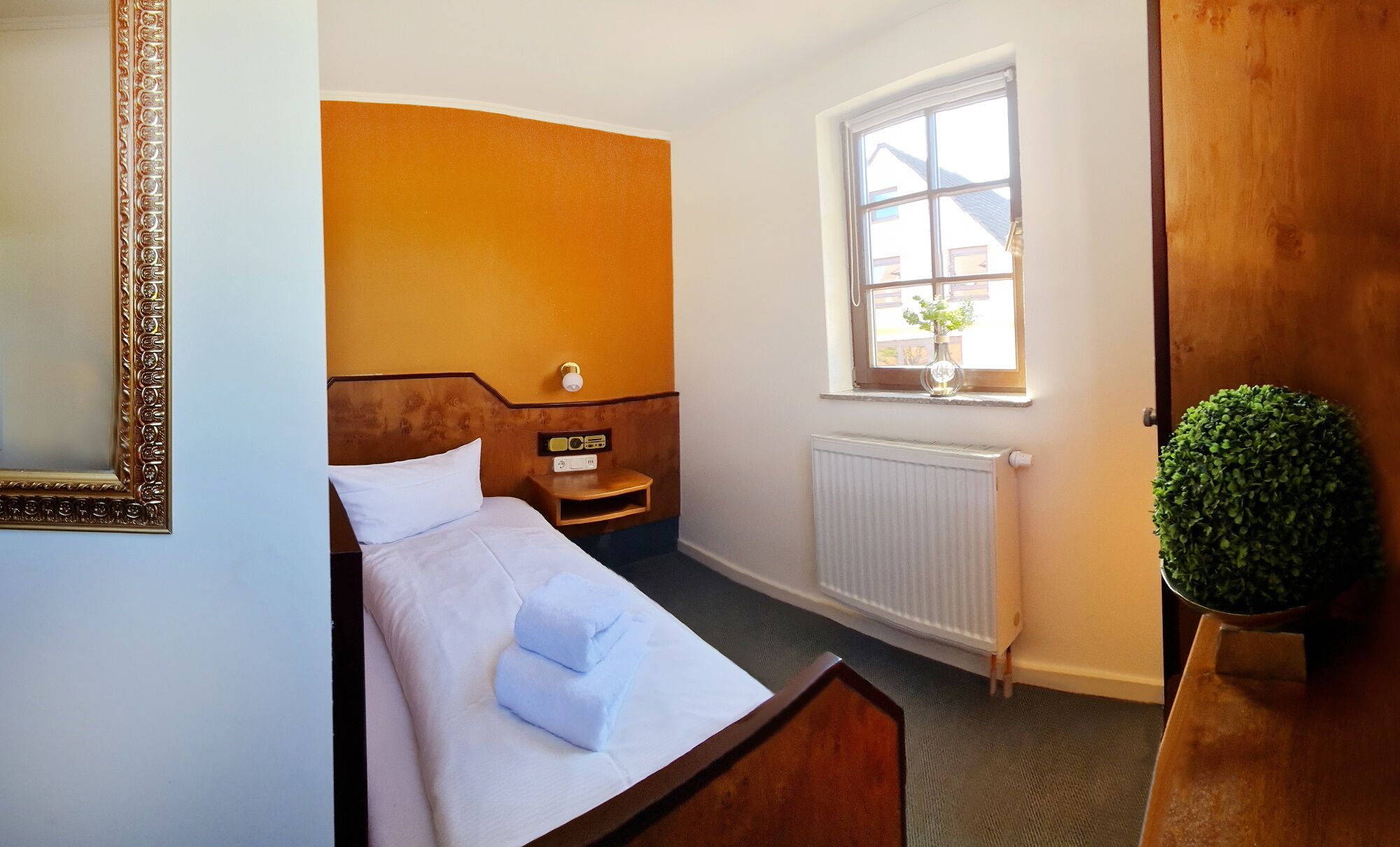 Ein Einzelbett mit weißem Kopfkissen und zusammengelegtem Handtuch, ein Nachttischchen mit einem Radio, ein Fenster mit Blick auf Gebäude und eine Heizung.