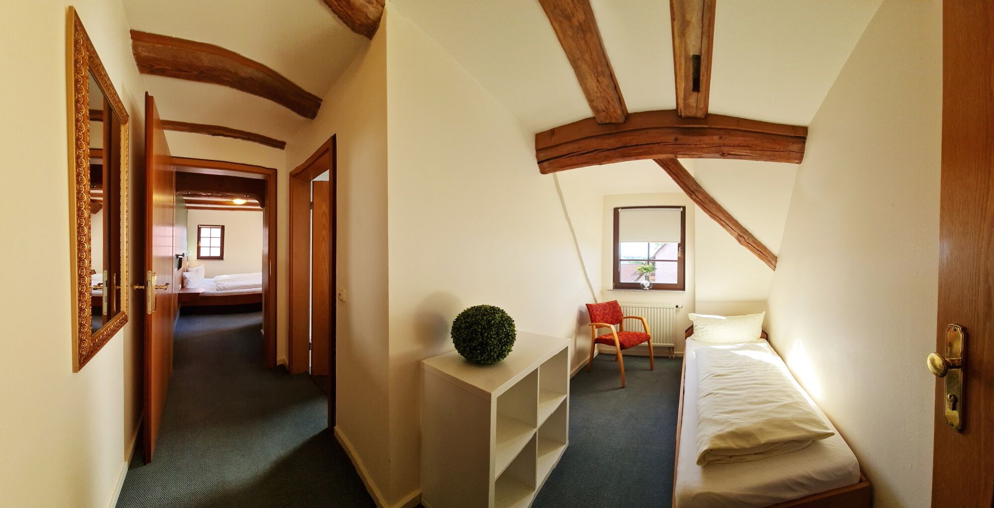 Een smalle gang die leidt naar een slaapkamer met een bed, een kleine tafel met een plant en een raam met uitzicht.