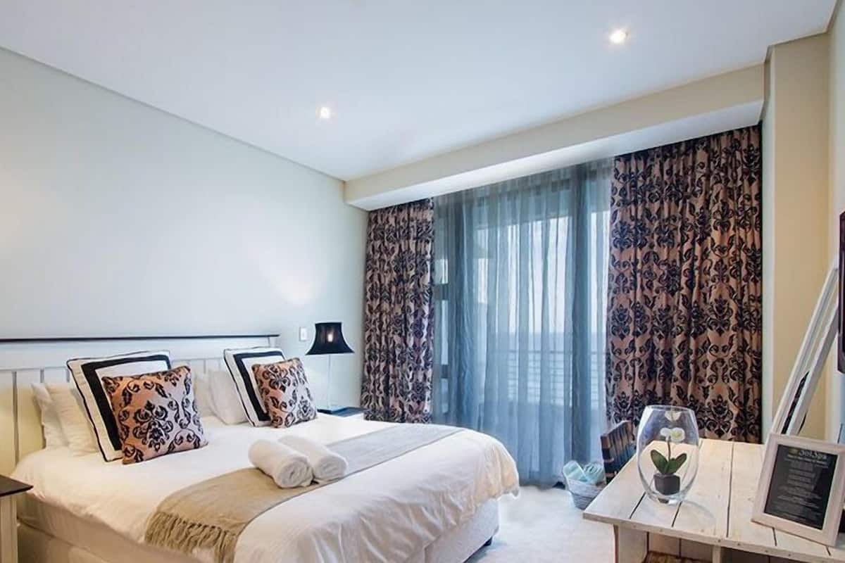 A hotel room with a large bed, two bedside tables, a desk with a chair, and a large window with curtains.