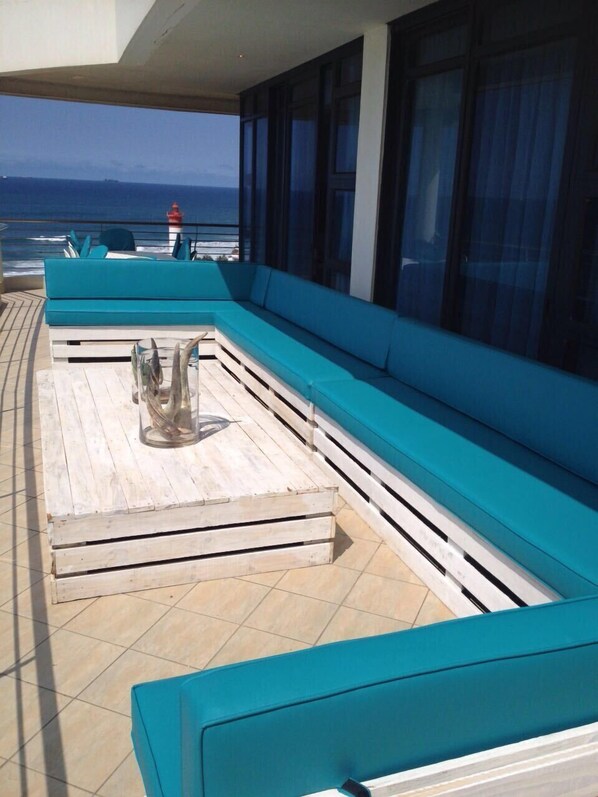 A sunlit outdoor seating area with a wooden table and blue cushioned benches, overlooking the sea.