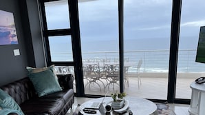 Ein modernes Wohnzimmer mit Ledersofa, hockerartigen Sesseln in Türkis und einem runden Couchtisch. Große Fenster bieten Blick auf Strand und Meer.