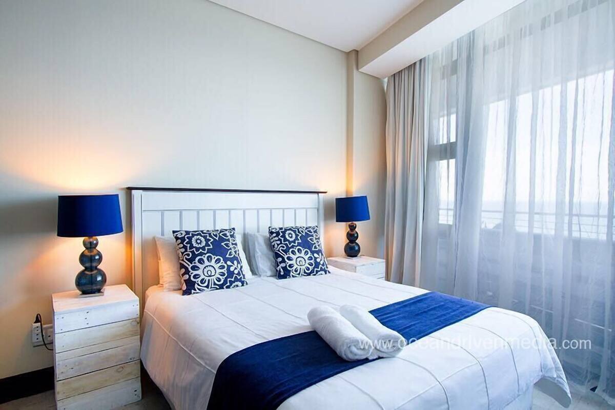 A hotel room with a bed, two bedside tables, lamps, and a large window with sheer curtains.