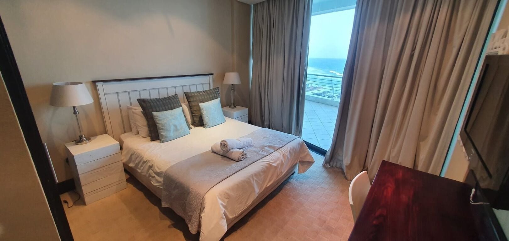 Quarto de hotel com cama, duas mesas de cabeceira, televisão e vista para o oceano através de amplas janelas.
