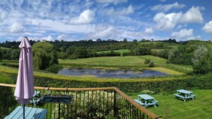 A scenic outdoor area with a wooden deck, a pond, and picnic tables. - Cameley Lodge (Bristol)