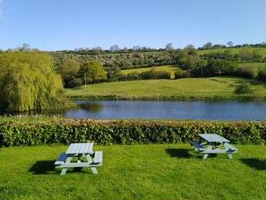 A serene lakeside setting with two picnic tables and a hedge in the foreground. - Cameley Lodge (Bristol)