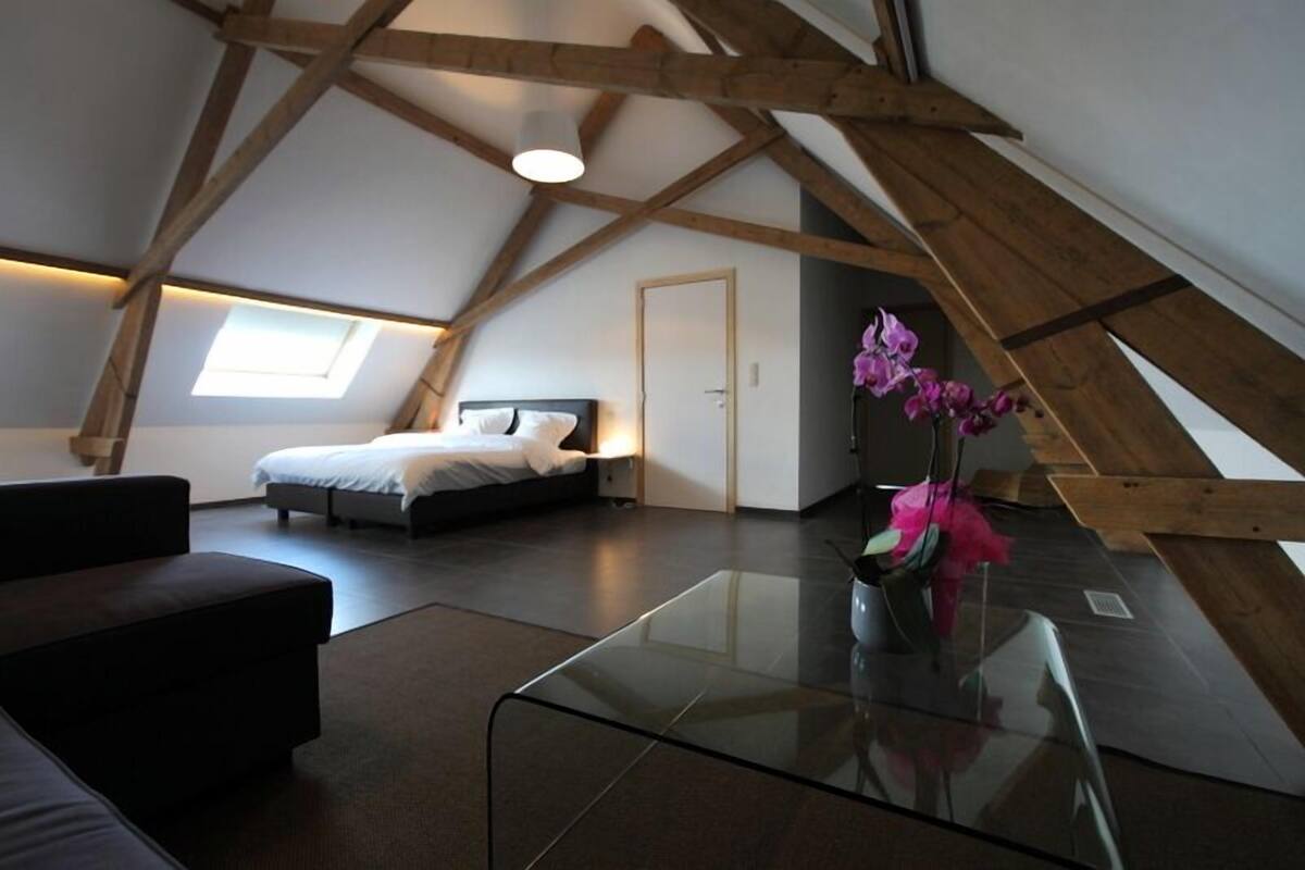 A modern bedroom with a sloped ceiling, a bed, a glass table, and a small window.