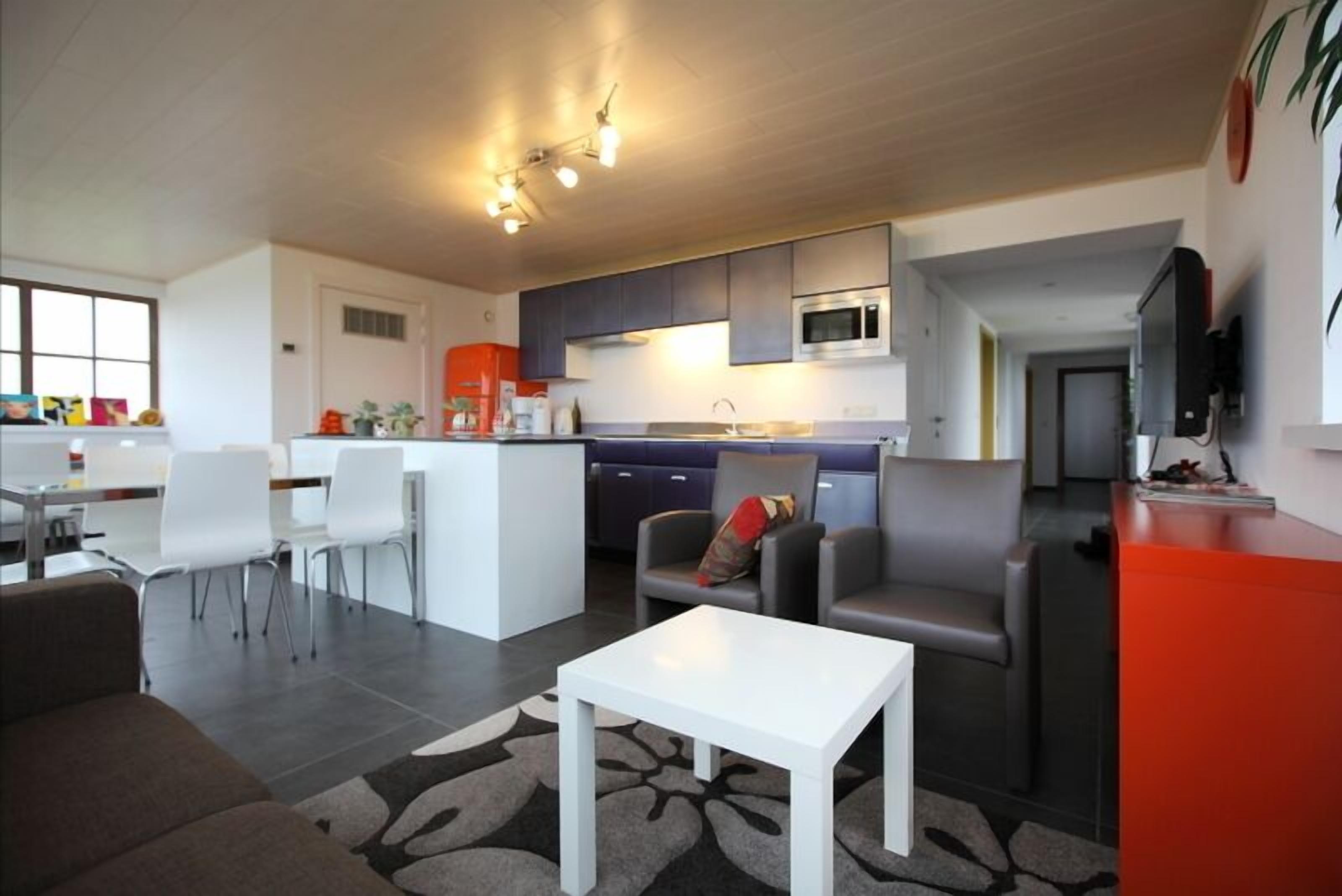 Een moderne woonkamer met een witte salontafel, grijze banken en een keuken met donkere kasten en een magnetron.