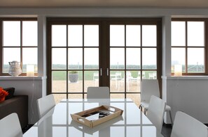 A modern dining area with a large table and chairs, large windows with a view of the outdoors, and a potted plant on the windowsill. - B&B Back2Front (Ypres)