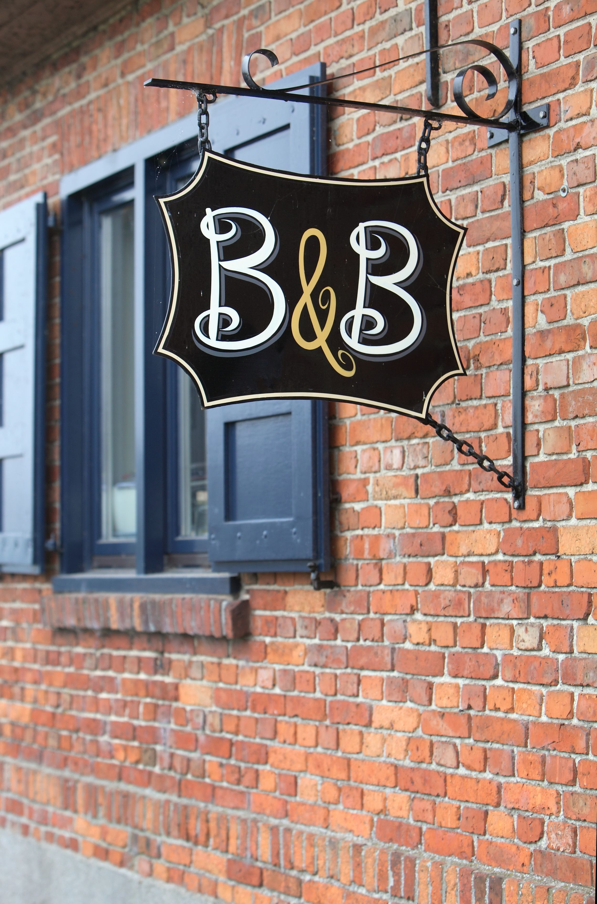 A black sign with white and gold lettering spelling "B&B" hanging on a brick wall.