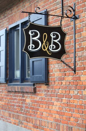 A black sign with white and gold lettering spelling "B&B" hanging on a brick wall. - B&B Back2Front (Ypres)