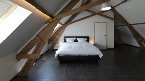 A bedroom with a sloped ceiling, exposed wooden beams, a bed with white bedding, and a door. - B&B Back2Front (Ypres)