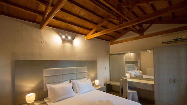 A hotel room with a wooden ceiling, a large bed with white linens, a bedside table with a lamp, a mirror, and a wardrobe. - Vicky Studios (Lesvos)