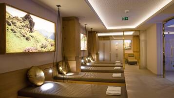 Una stanza spa con diversi lettini per massaggi, pannelli in legno e un grande schermo a parete che mostra un paesaggio.