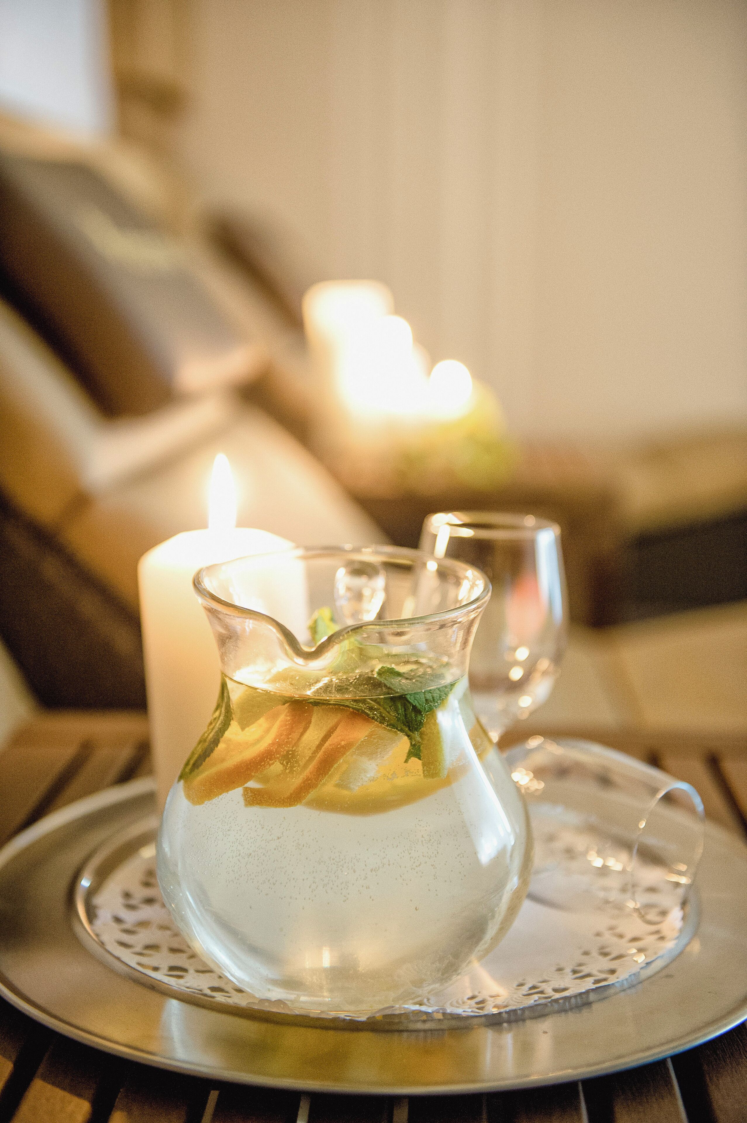 A clear glass pitcher with a beverage and mint leaves on a tray, accompanied by a lit candle and a glass on a wooden table.