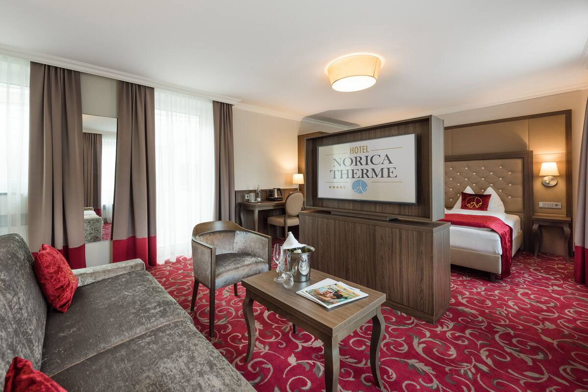 A hotel room with a sofa, a small table, a TV displaying the hotel name, a bed, and a red carpet.