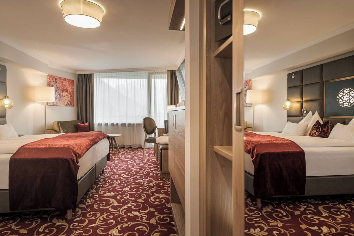 A hotel room with two beds, a desk, a chair, and a large window with curtains.