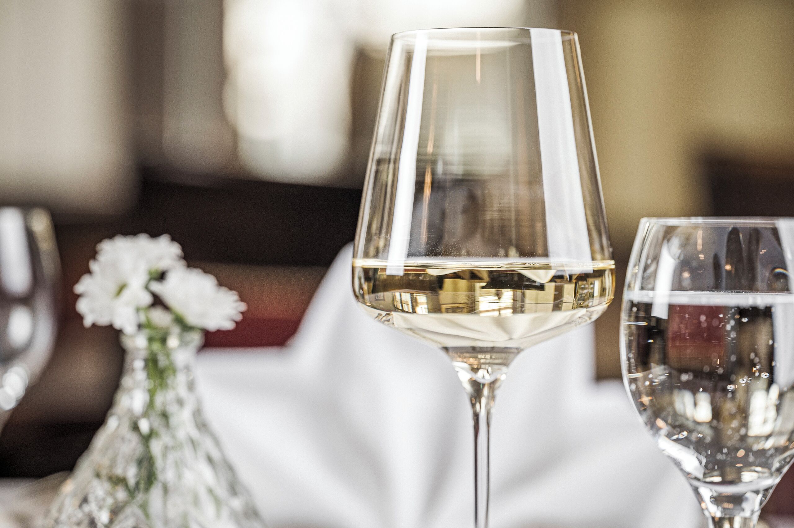 A close-up of a wine glass with a clear liquid, a smaller glass with a clear liquid, and a white vase with white flowers on a table.