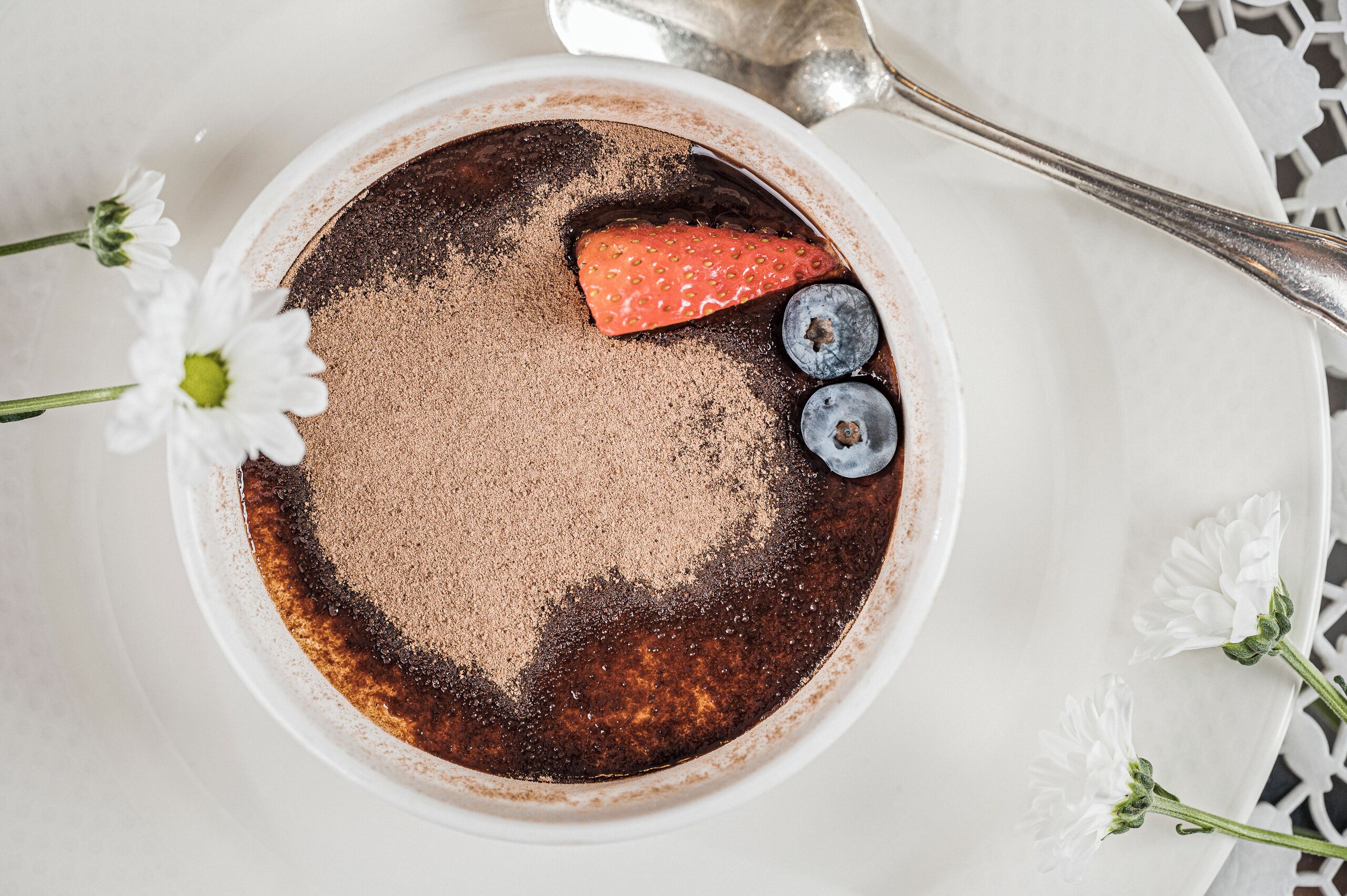 A dessert in a white bowl with a strawberry and blueberries on top, surrounded by white flowers.