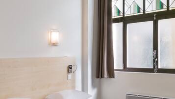 Une chambre d’hôtel avec une tête de lit en bois, deux lits avec une literie marron et blanche, un lampadaire fixé au mur, une fenêtre ornée d’un motif géométrique et un radiateur.