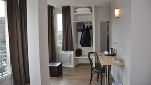 A compact hotel room with a wooden floor, a small table with a chair, a radiator, a coat rack with jackets, and a window with curtains. - Hotel Foch Nancy Centre Gare (Nancy)