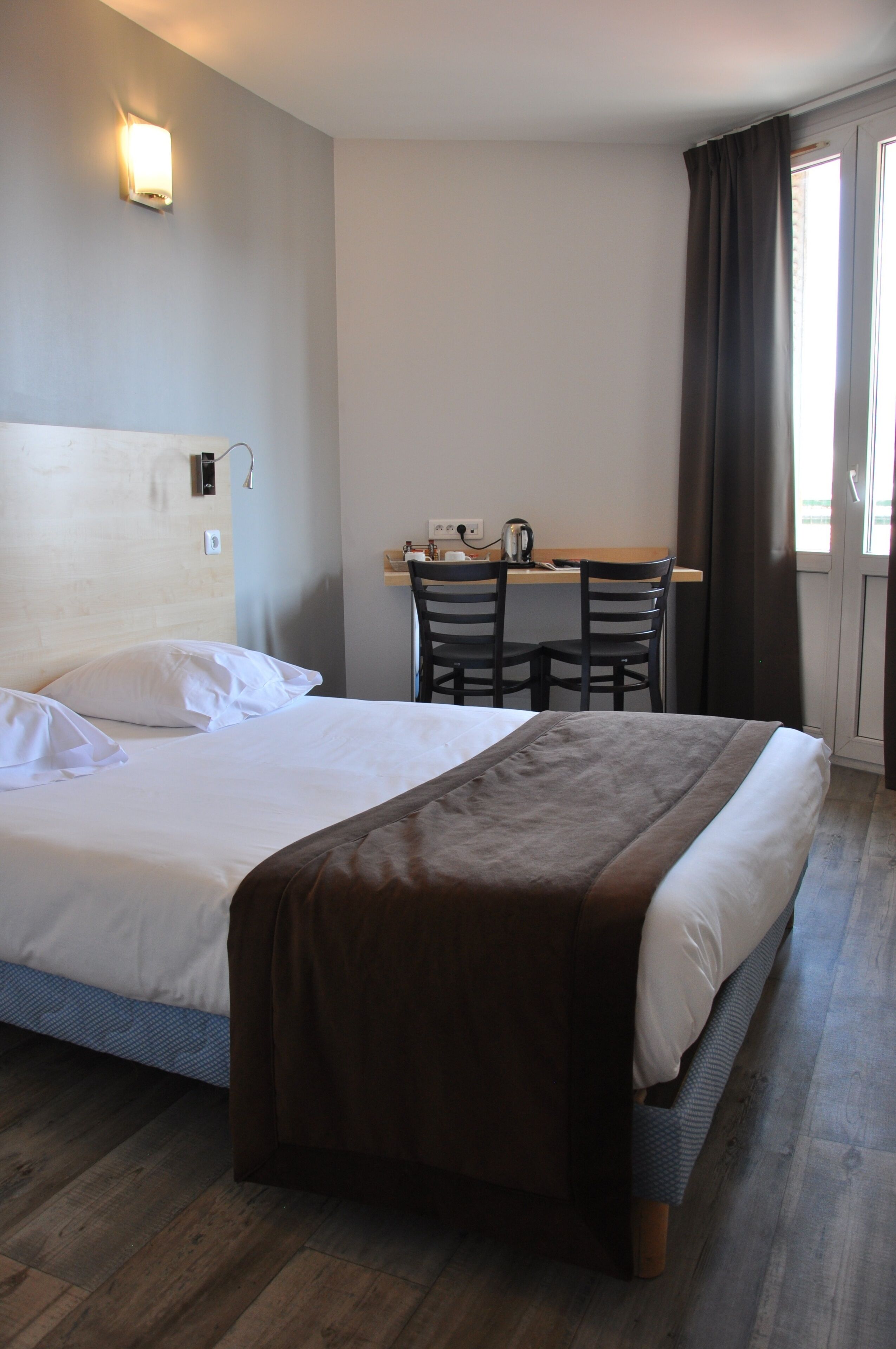 A hotel room with a bed, a desk with a kettle, and a window with curtains.