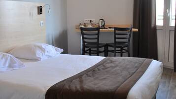 A hotel room with a bed, a desk with a kettle, and a window with curtains.