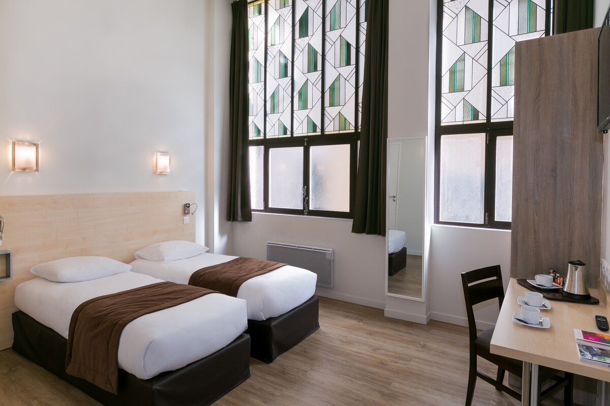 A hotel room with two beds, a desk with a chair, a radiator, and a large window with curtains.