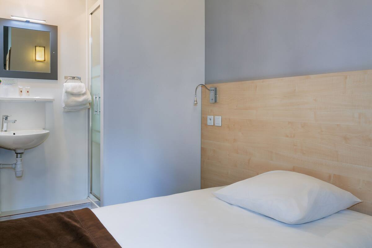 A hotel room with a bed, a white pillow, a wooden headboard, a white sink, and a glass shower enclosure.