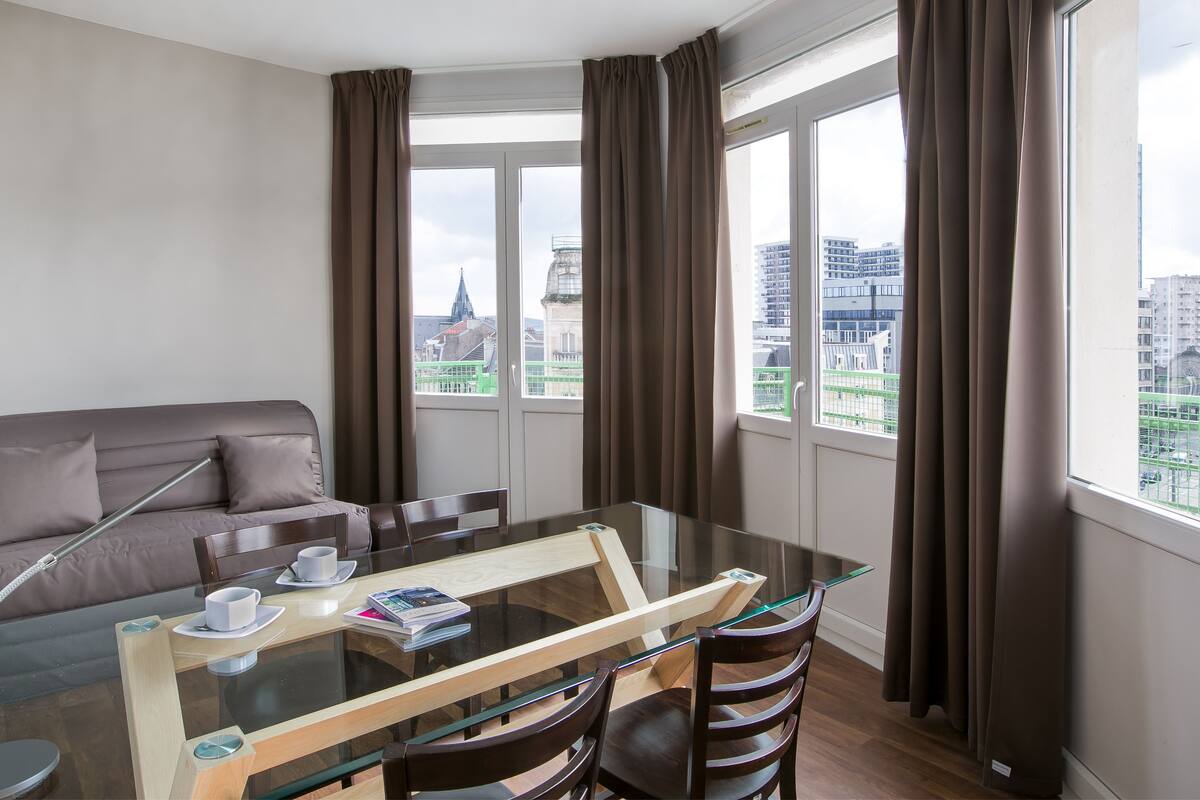 A modern hotel room with a glass dining table, brown leather sofa, and large windows offering city views.