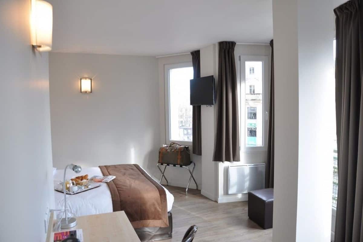 A hotel room with a bed, bedside table, lamp, TV, and a window with curtains.