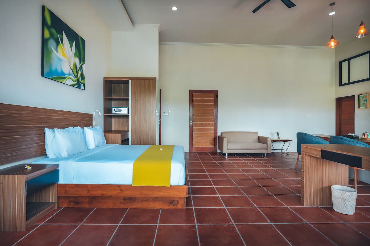A hotel room with a bed, a desk, a chair, a sofa, a wooden wardrobe, and a painting on the wall.
