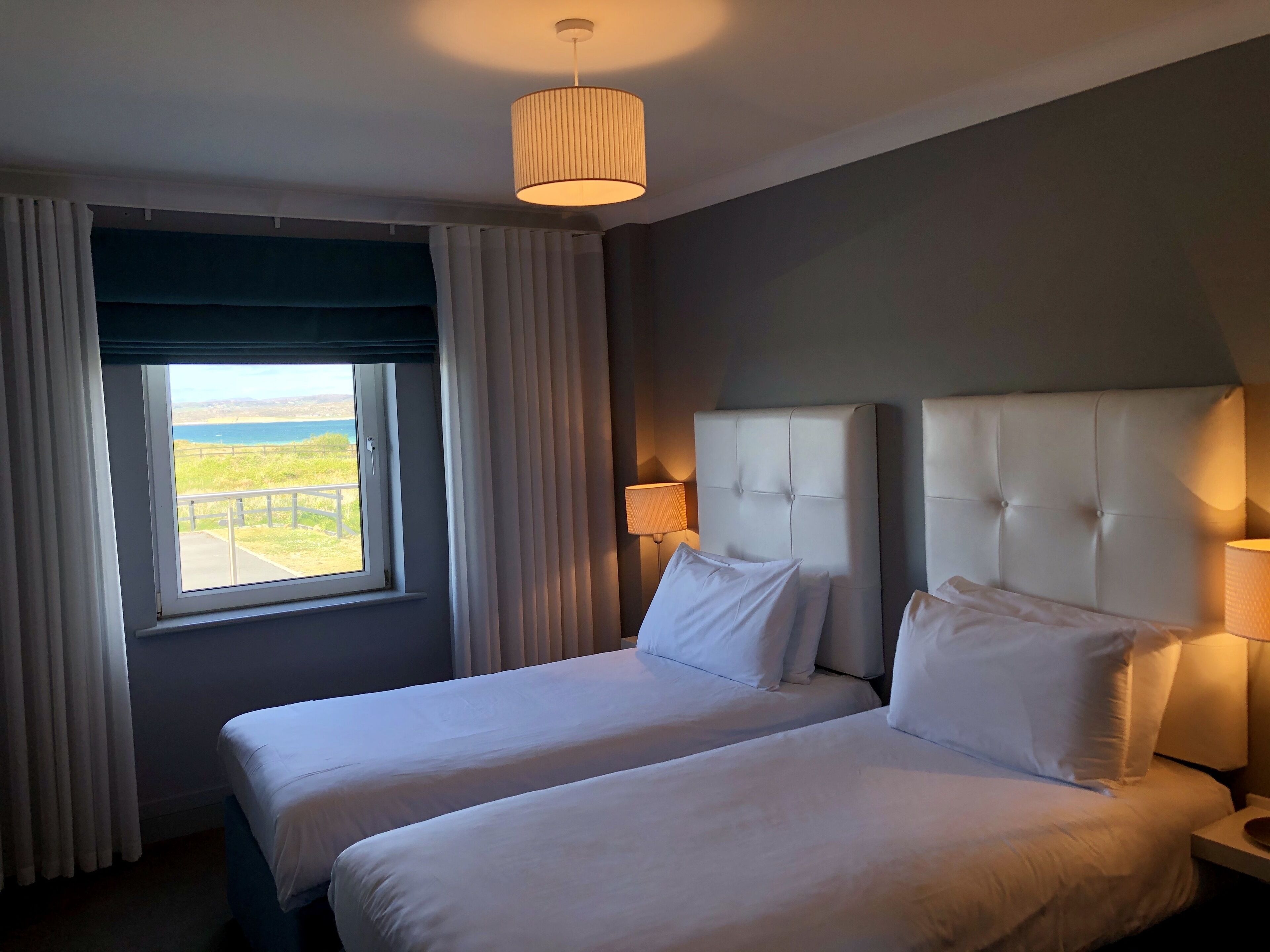 Une chambre d’hôtel avec deux lits, une grande fenêtre avec des rideaux, un luminaire suspendu et une vue sur l’extérieur.