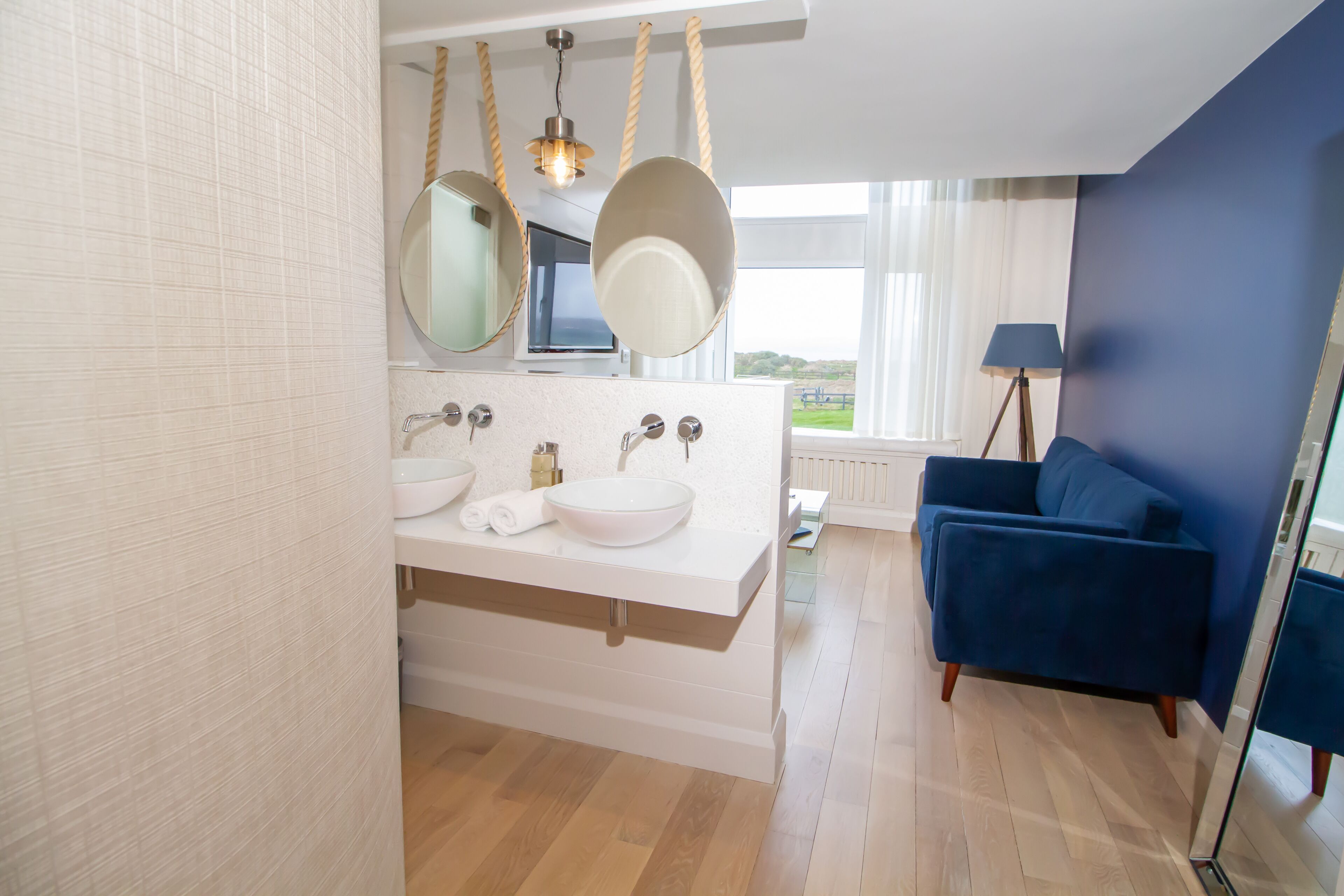 Un bagno moderno con un mobile lavabo bianco, due specchi e un divano blu nella zona soggiorno adiacente.