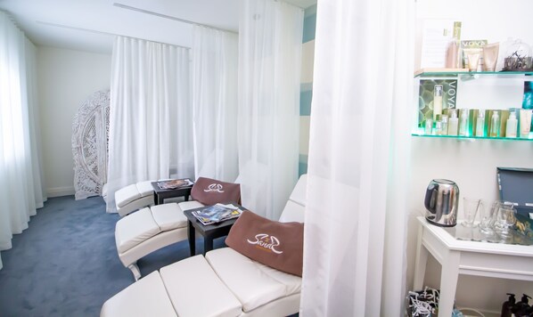 Una habitación moderna con repisas de vidrio exhibiendo artículos de aseo, una cama blanca con dosel y una zona de estar con una revista y un cojín.