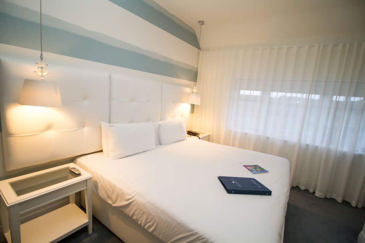 A hotel room with a large bed, two bedside lamps, a nightstand, and a window with sheer curtains.