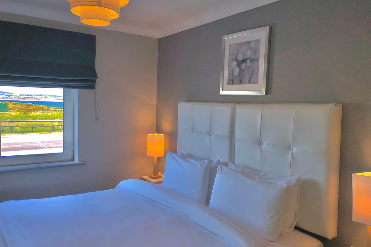 A bedroom with a large bed, a window with a view of greenery and mountains, a framed picture on the wall, and a bedside lamp.