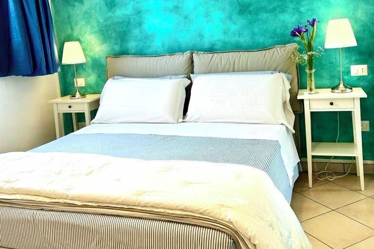 Ein Bett mit blau-weiß gestreifter Bettdecke, zwei weiße Nachttische mit Lampen und eine wandfarbene Wand in Türkis.