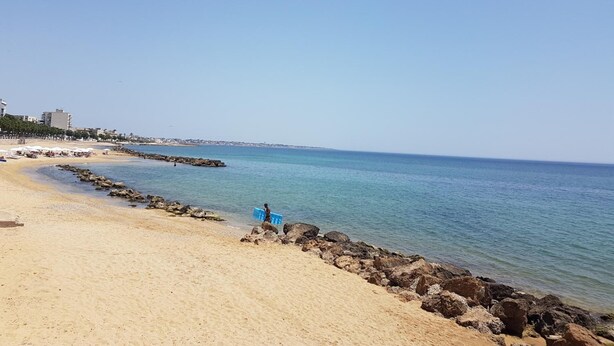 Ein Strand mit einem steinernen Wellenbrecher, klarem blauen Wasser und Gebäuden im Hintergrund.