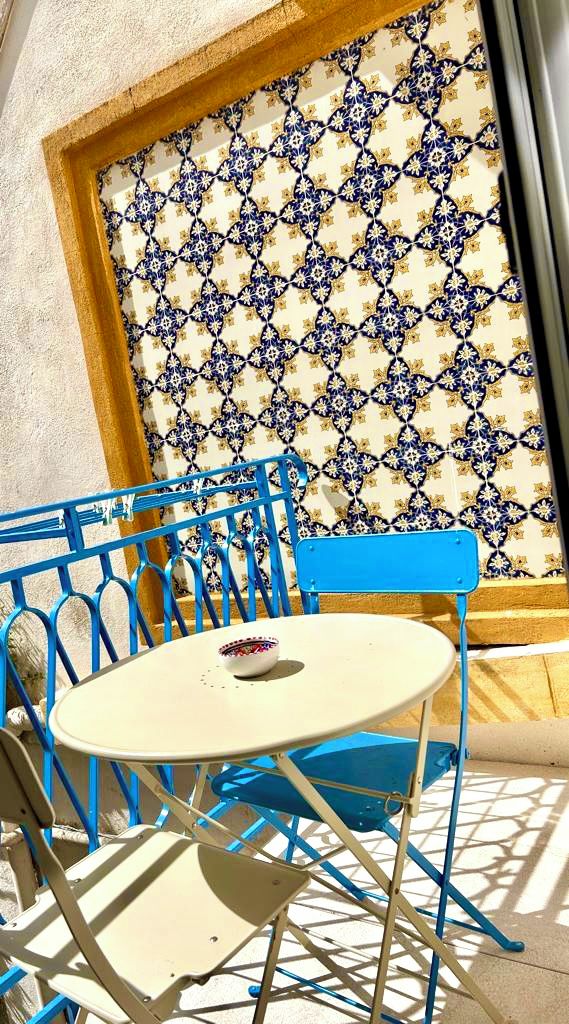 A sunlit outdoor seating area with blue metal chairs and a white table, featuring a patterned wall in the background.