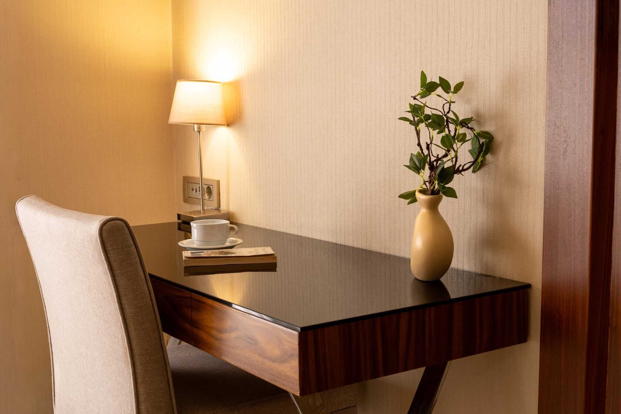 A hotel room with a wooden desk, a white chair, a bedside lamp, a small potted plant, and a cup on the table.