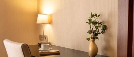 A hotel room with a wooden desk, a white chair, a bedside lamp, a small potted plant, and a cup on the table.