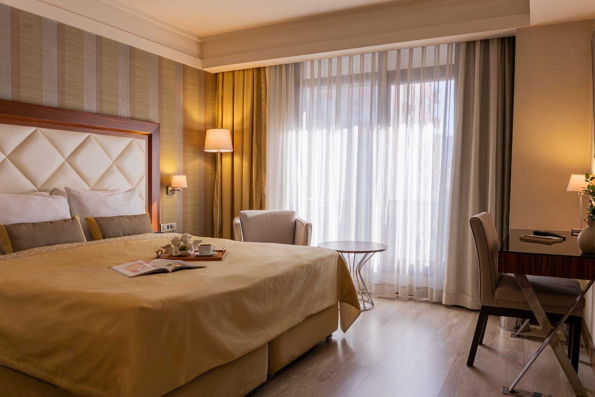 A hotel room with a large bed, a desk, a chair, a small table, and a window with curtains.