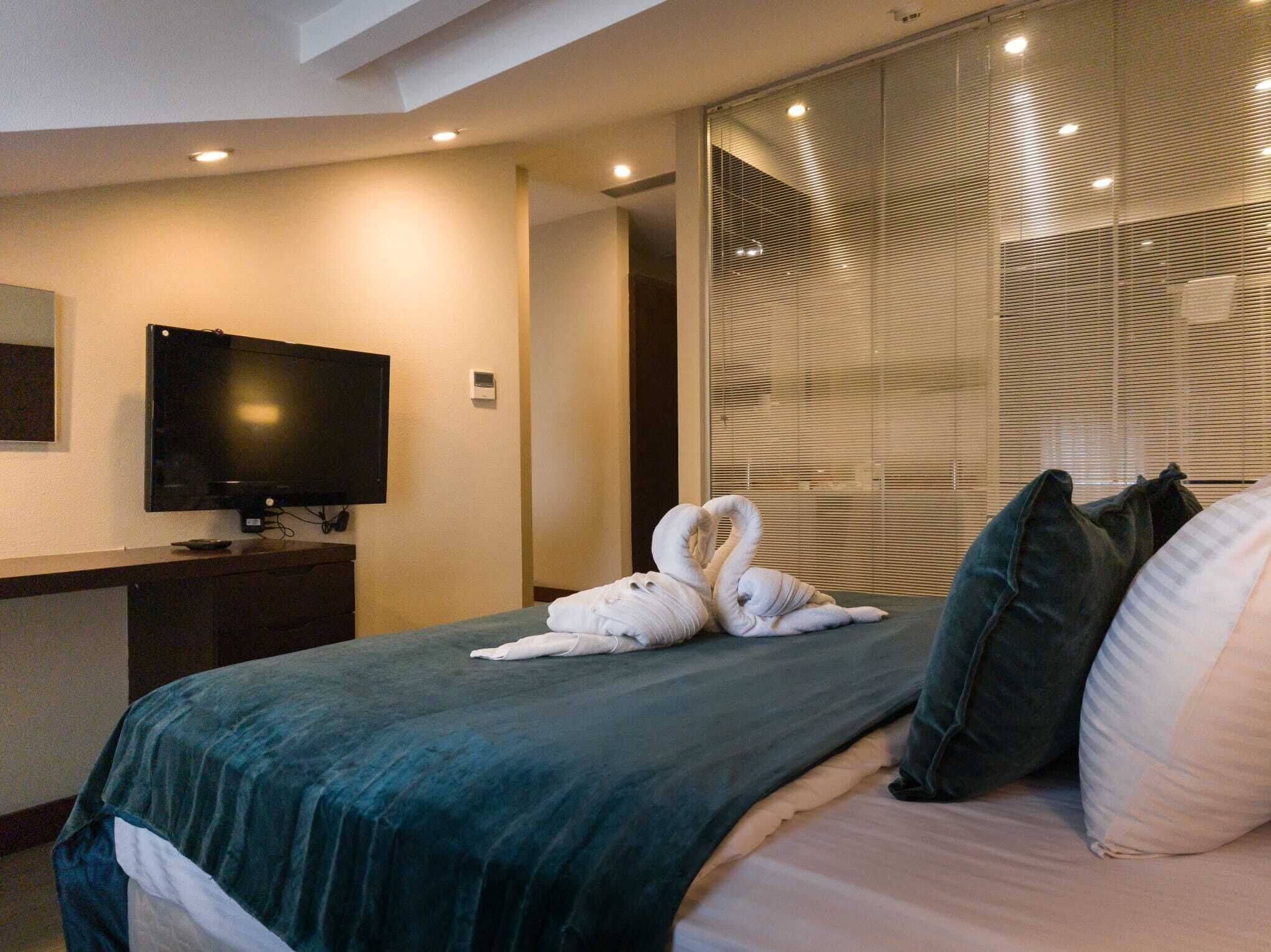 A hotel room with a bed, a flat-screen TV, a desk, and a large window with blinds.