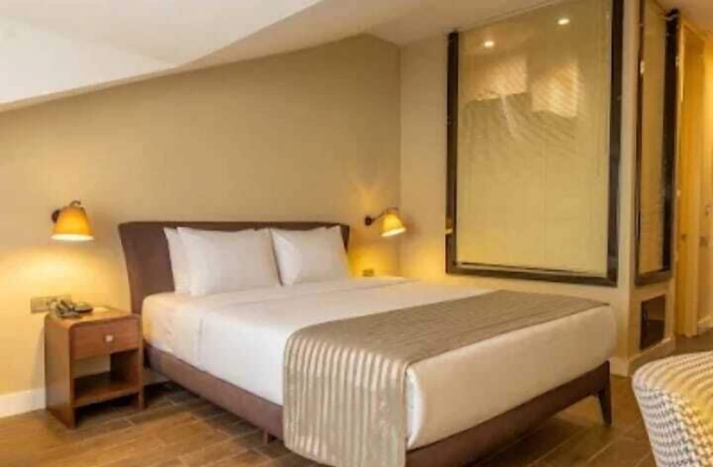 Habitación de hotel con una cama grande, lámparas de noche, una mesita de noche y una ventana con persianas.
