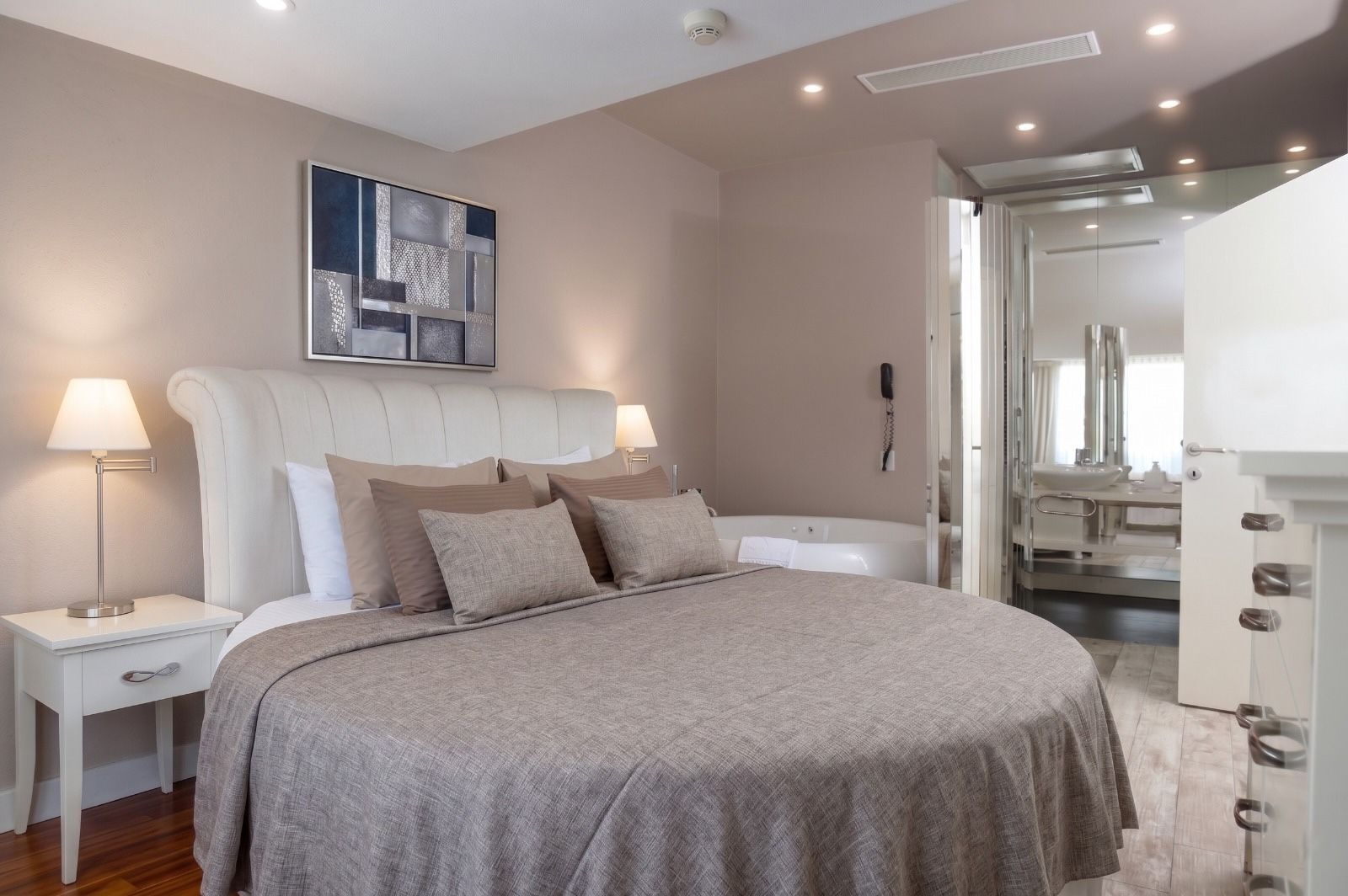 Un dormitorio con una cama grande, mesitas de noche, cabecero, una obra de arte colgada en la pared, y un baño visible a través de una puerta abierta.