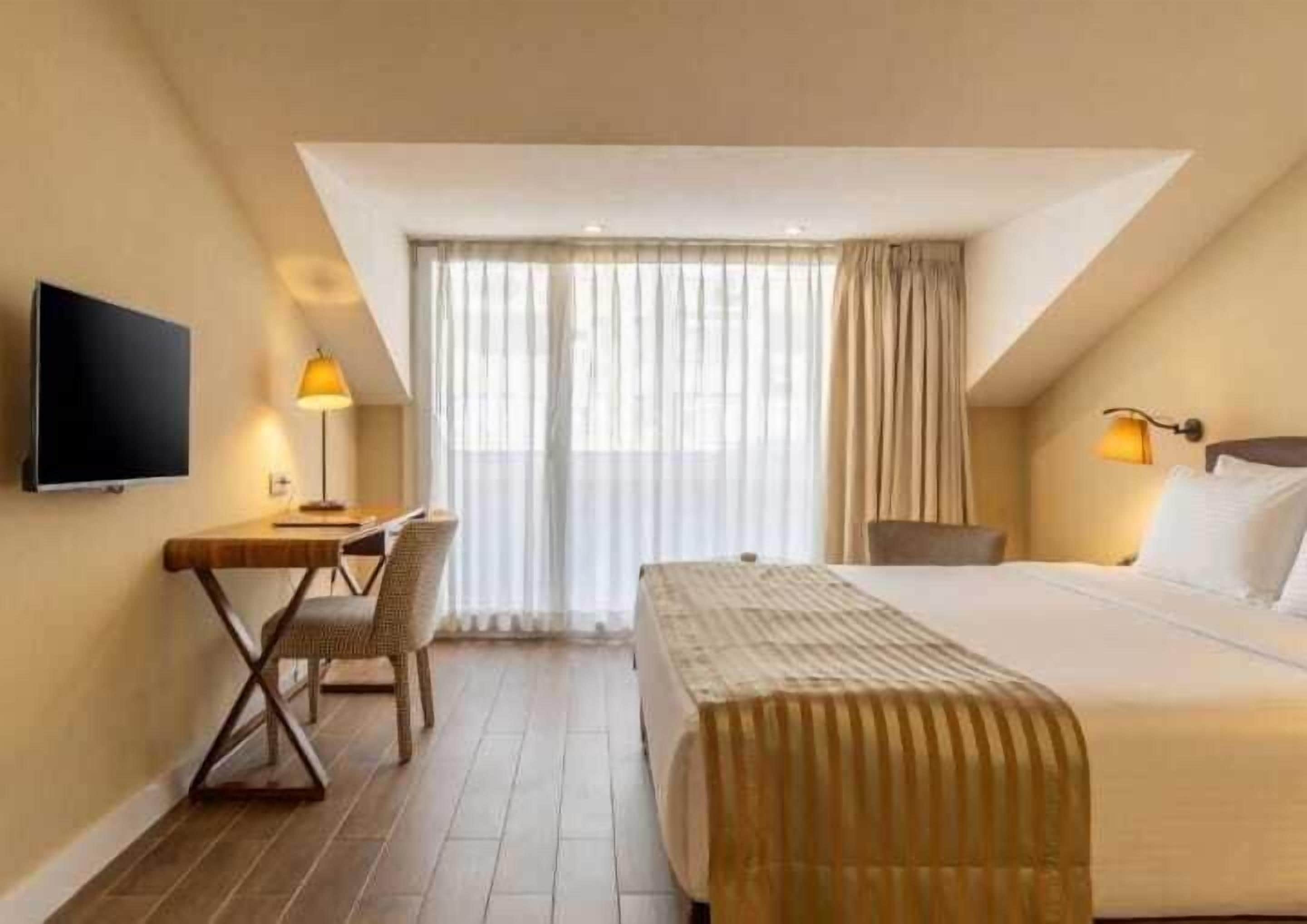 Una habitación de hotel con cama, escritorio, silla, televisor y ventana con cortinas.