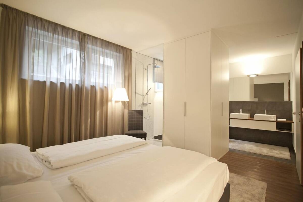 A hotel room with a bed, a chair, a bathroom with a mirror and sinks.