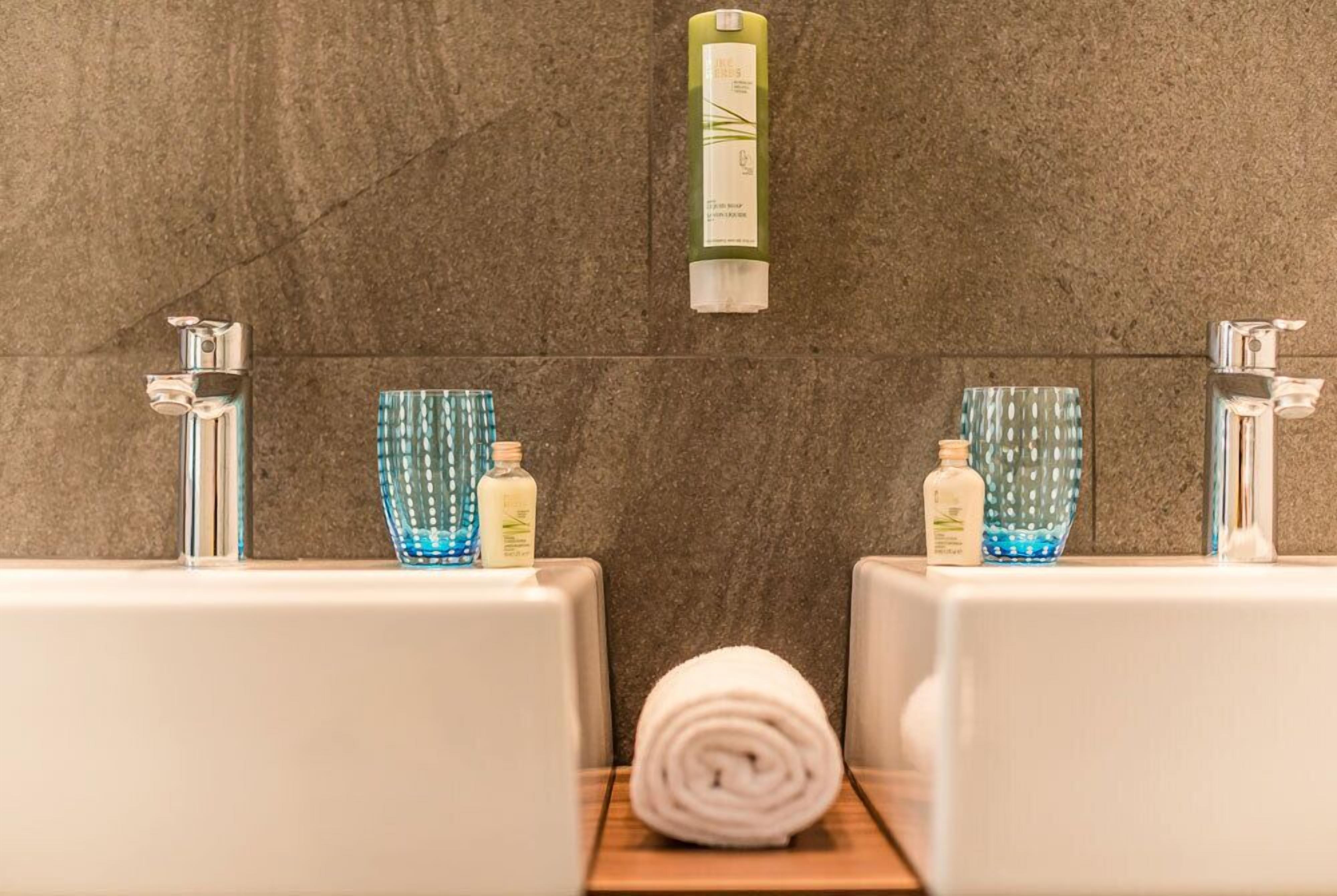 A bathroom with two sinks, each with a bottle of soap and a blue glass container. A towel is rolled up in the center.