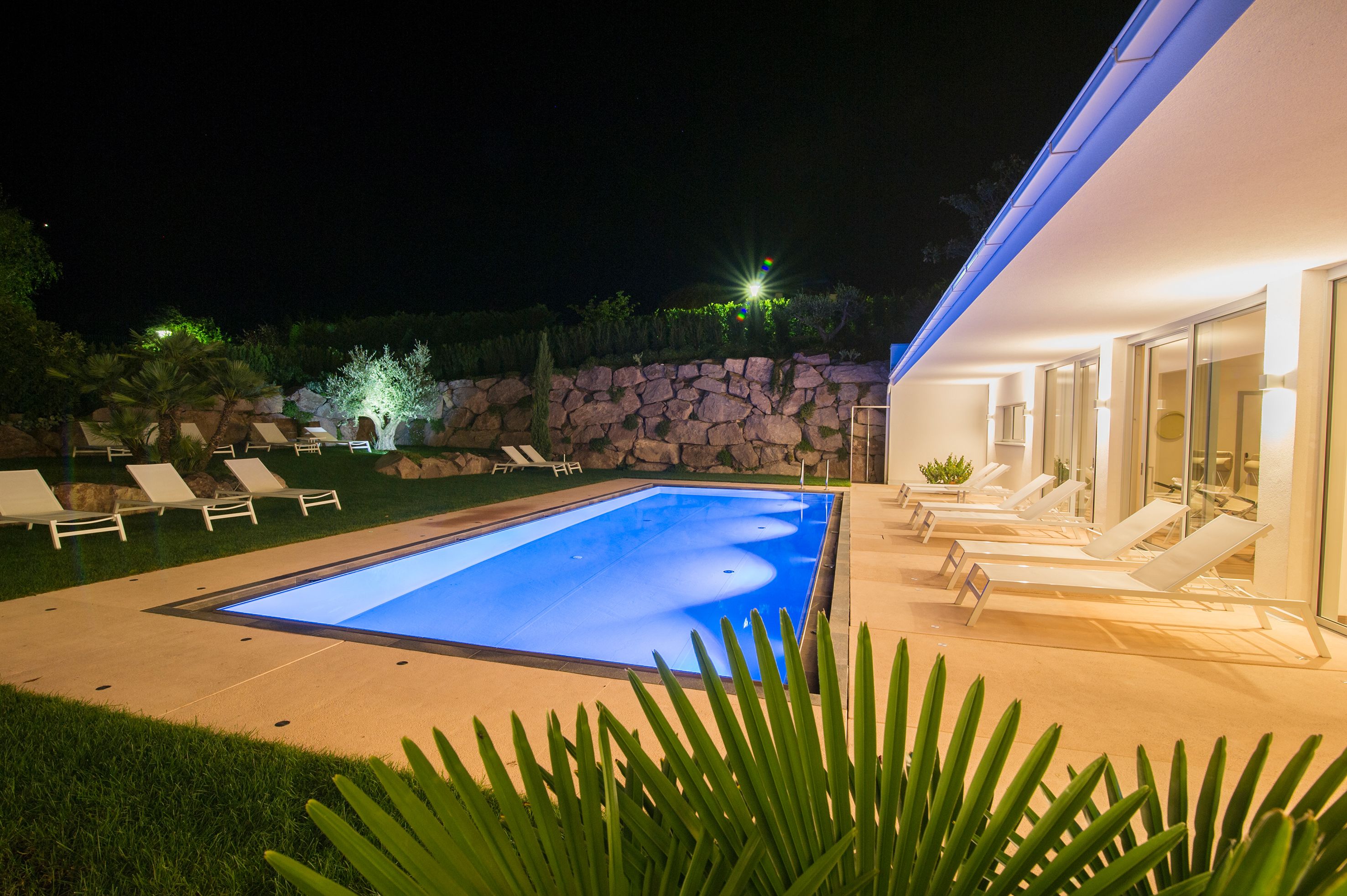 En swimmingpool med solstolar, en stenmur och en modern byggnad med glasdörrar.