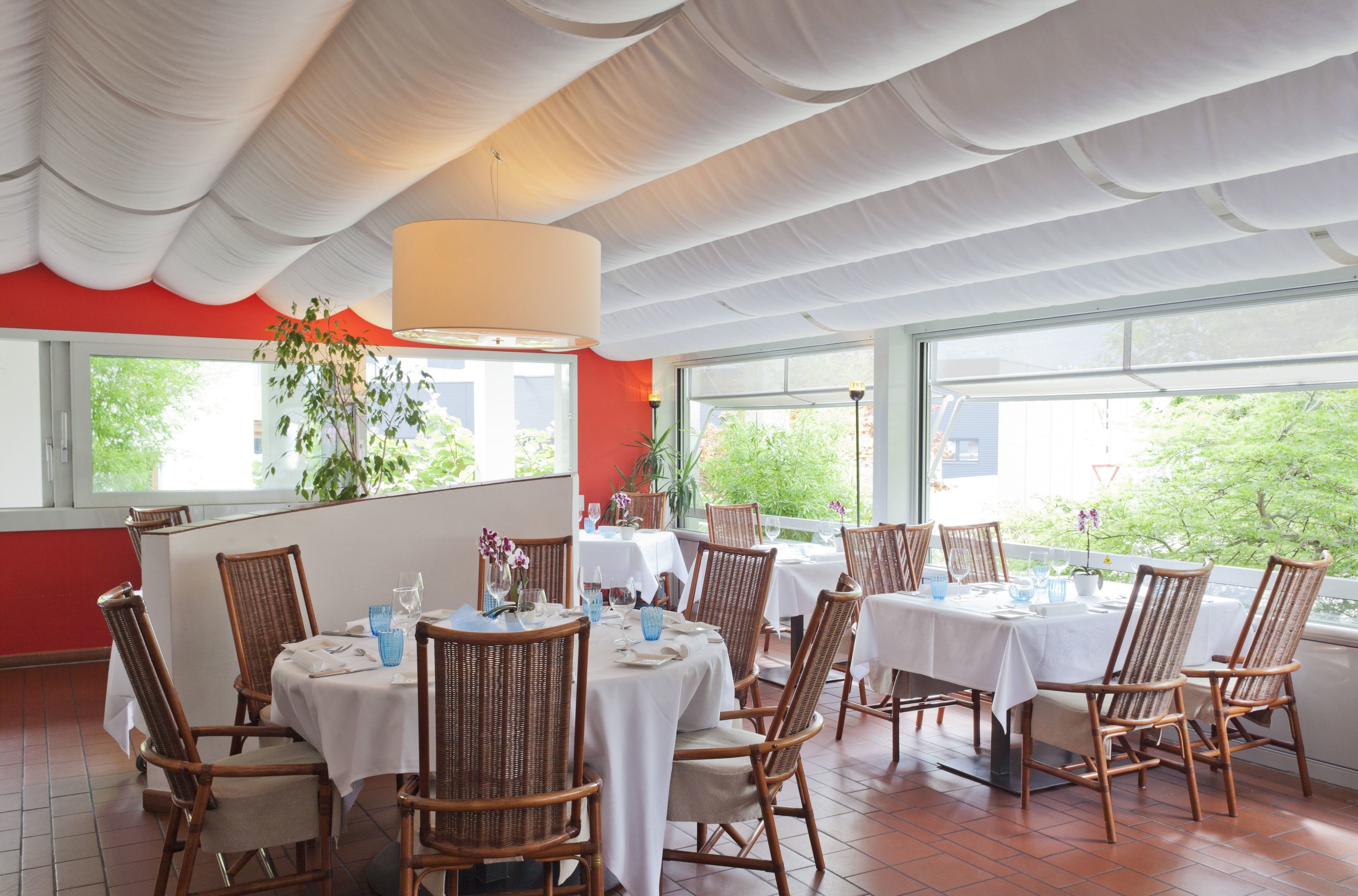 Un restaurante con varias mesas, sillas de mimbre, ventanas amplias y un techo blanco.